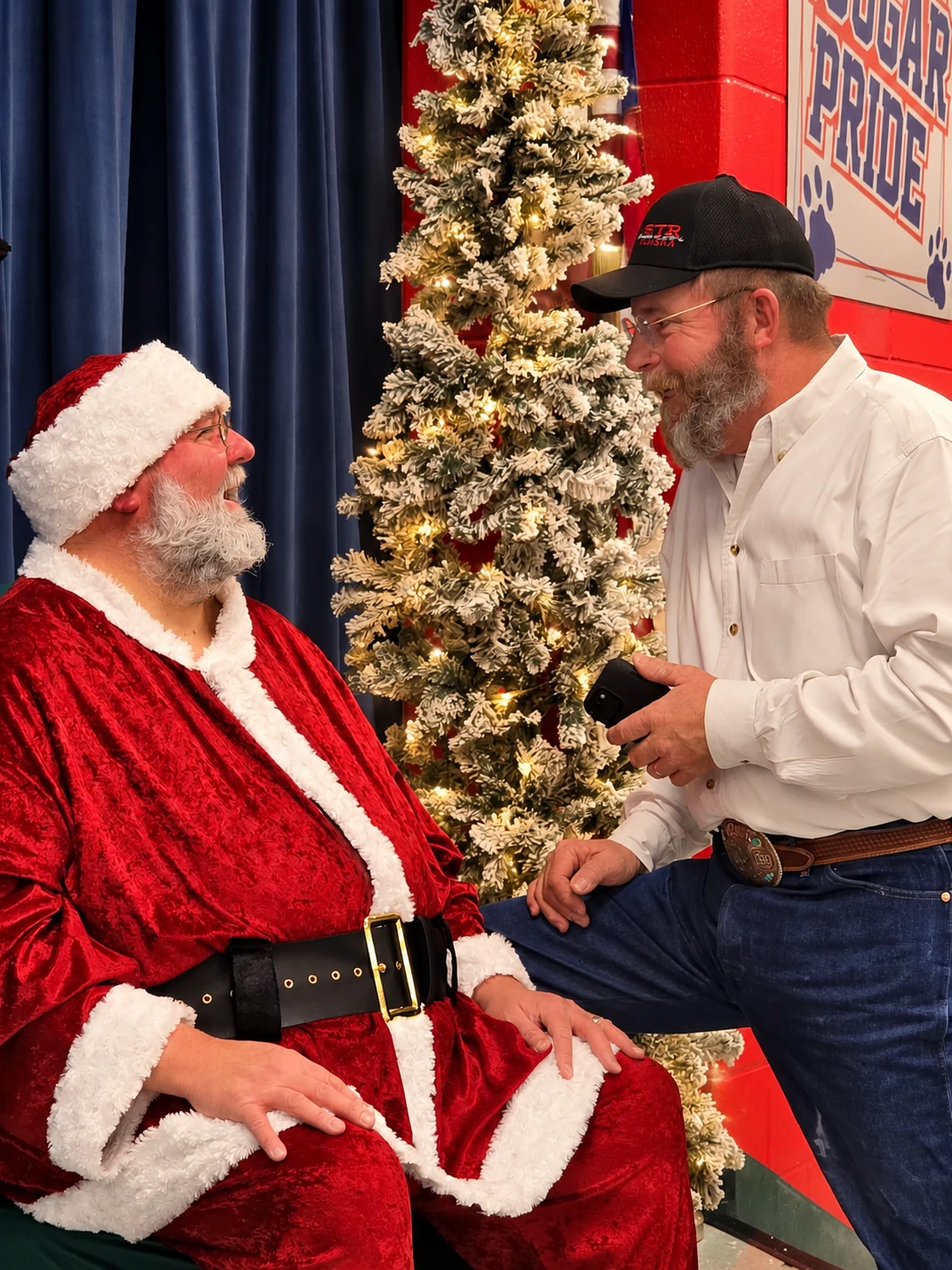 Santa Jeff sharing a conversation with a guest beside a lit Christmas tree and a Cougar Pride sign