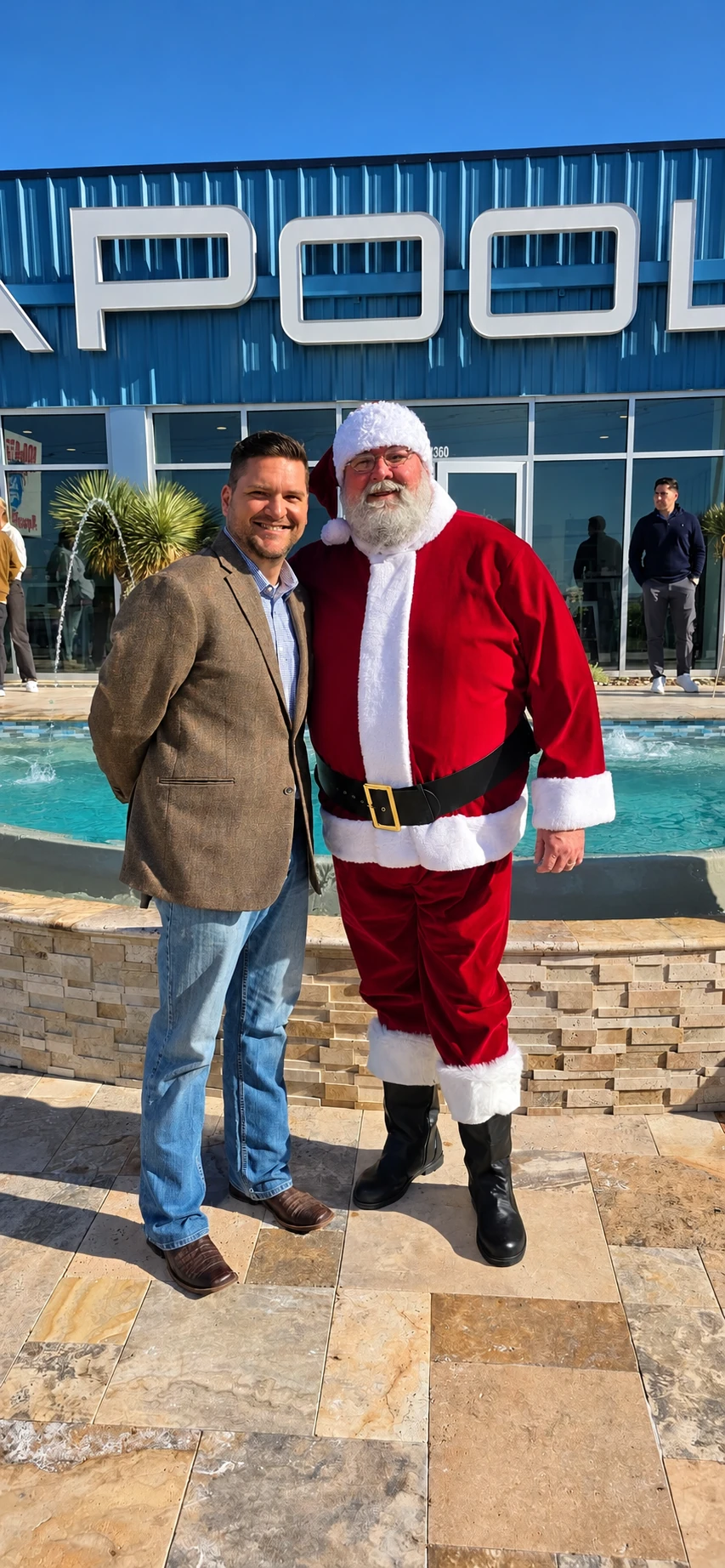 Santa Jeff and a smiling guest standing beside an outdoor fountain on a sunny day